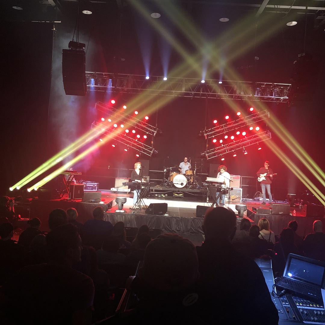A live band mid-set under crossing light beams, framed from the front-of-house mix position
