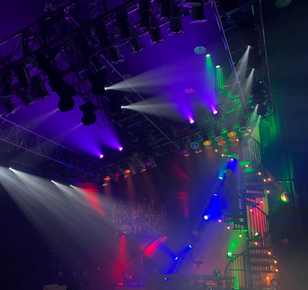 Concert lighting rig mid-program — trussed fixtures throwing purple and white beams across the stage